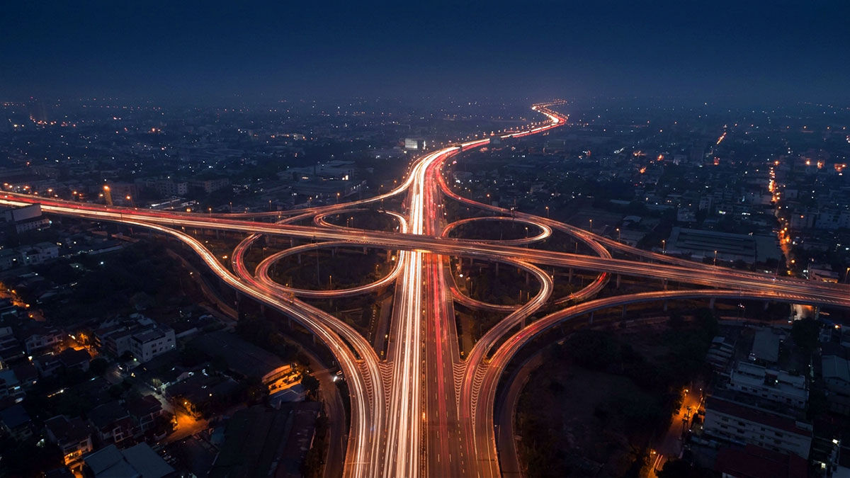 Aerial photograph of highway interchange at night with long-exposure light trails showing red and white streams of traffic forming branching patterns